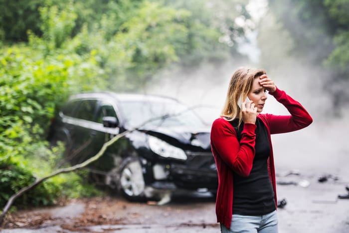 A young woman with smartphone by the damaged car after a car accident, making a phone call.