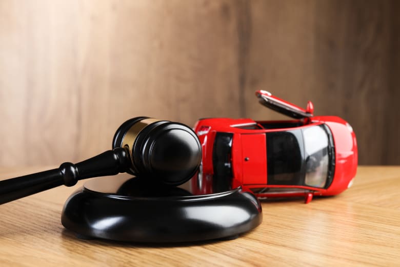 Mazo de juez de madera apoyado sobre un bloque de sonido frente a un automóvil rojo dañado después de un accidente, simbolizando la ley de accidentes automovilísticos y los procedimientos legales.