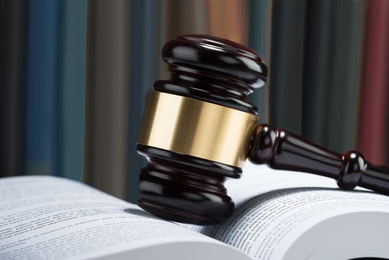 Close-up of a judge's wooden gavel with a brass band resting on an open law book, symbolizing legal authority and justice, with a blurred background of bookshelves.