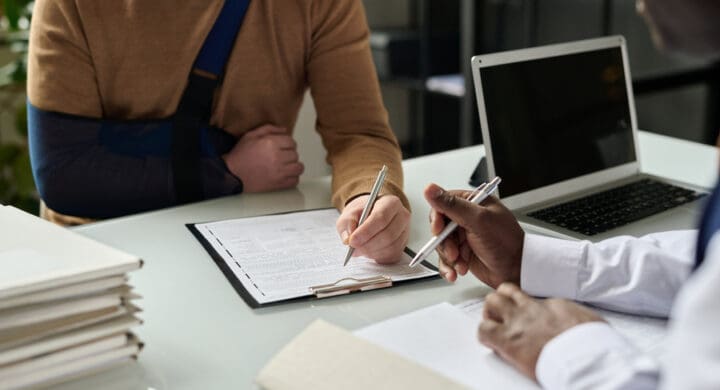 Close up of man filling in medical insurance form, injured hand, and discussing his personal injury case with lawyer