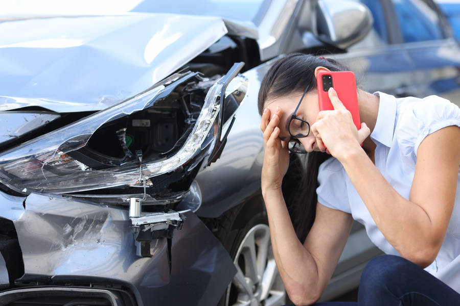 Mujer triste hablando por teléfono celular cerca de un auto averiado después de un accidente.